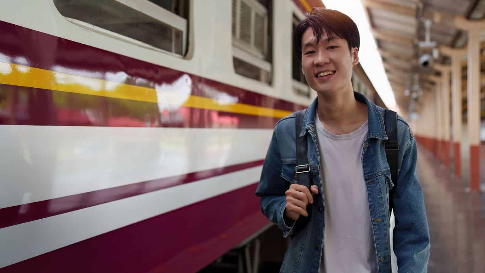 A young man wearing a denim jacket and backpack stands on a train platform next to a maroon and white train, smiling at the camera. The platform is mostly empty and covered by a roof.