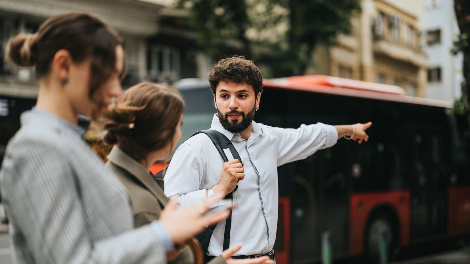 A man with a backpack points toward a red bus while talking to two women on a city street. The women are looking at their phones, and buildings are visible in the background.