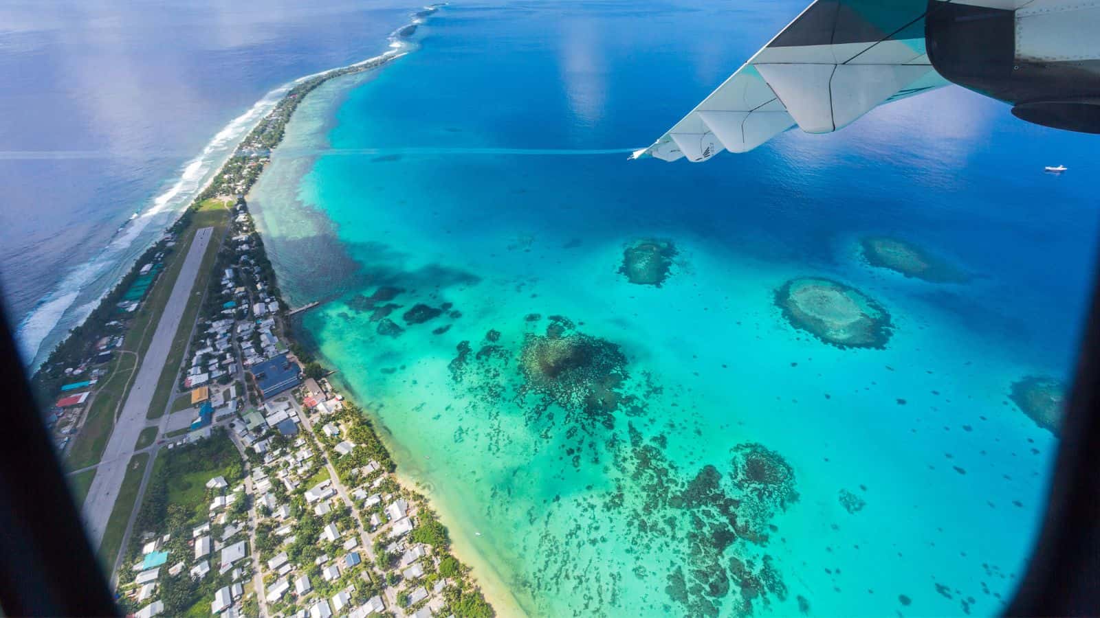 Aerial view from an airplane window showing an island with a runway, coastal buildings, turquoise water with coral reefs, and another airplane—an evocative glimpse of vanishing countries threatened by rising seas.