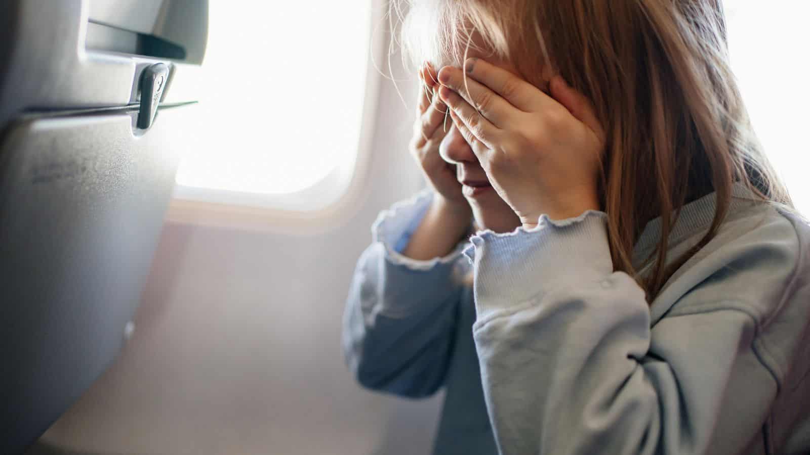 A child sits in an airplane seat next to a window, covering their face with both hands, perhaps trying to overcome flight anxiety. The child is wearing a light blue long-sleeved shirt as sunlight streams through the window.