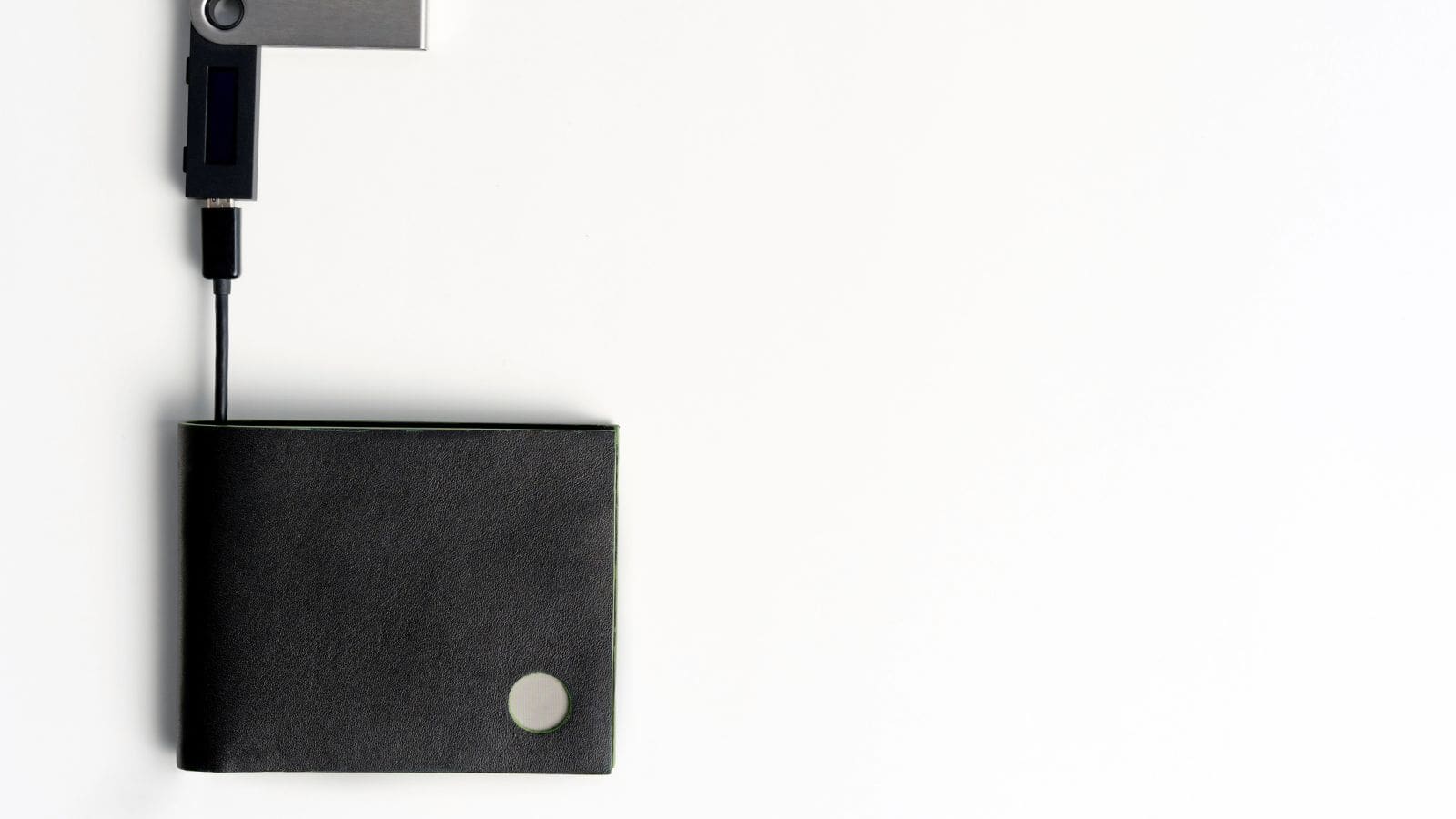A black wallet lies on a white surface with a cryptocurrency hardware wallet and a USB cable attached to it. The wallet is positioned at the lower left, while the devices are at the top left of the image.
