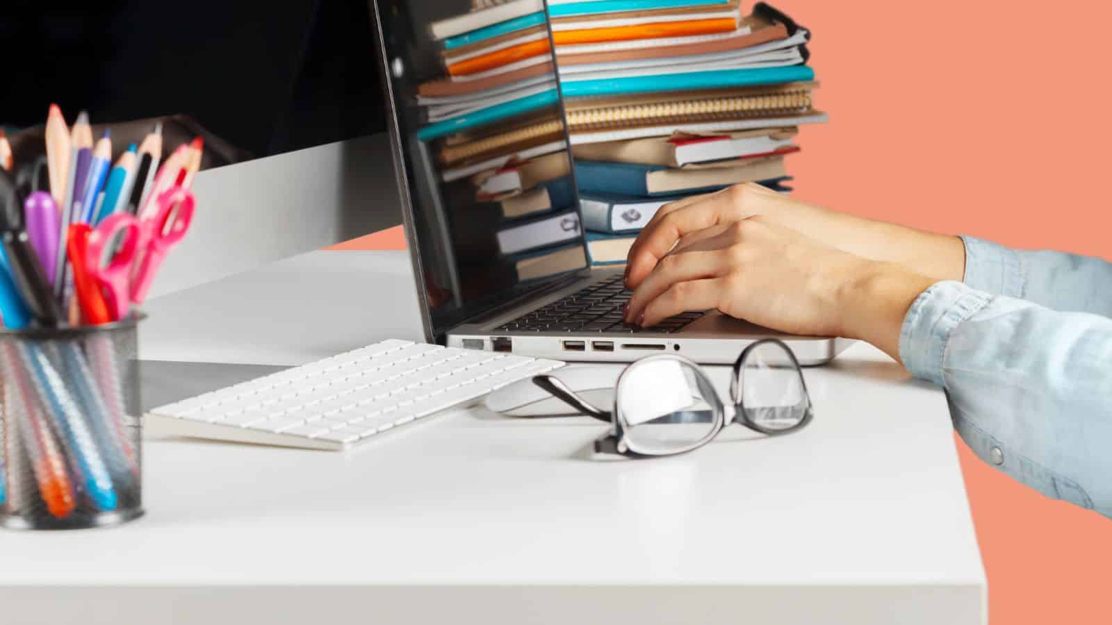 A person types on a laptop at a white desk with a desktop computer, eyeglasses, a cup of colored pencils, and a stack of notebooks and folders in the background.