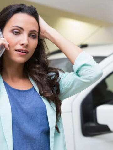 A woman stands in front of a white vehicle, holding a phone to her ear with one hand and touching her head with the other, appearing concerned or stressed. She is wearing a light blue blazer and a blue shirt.