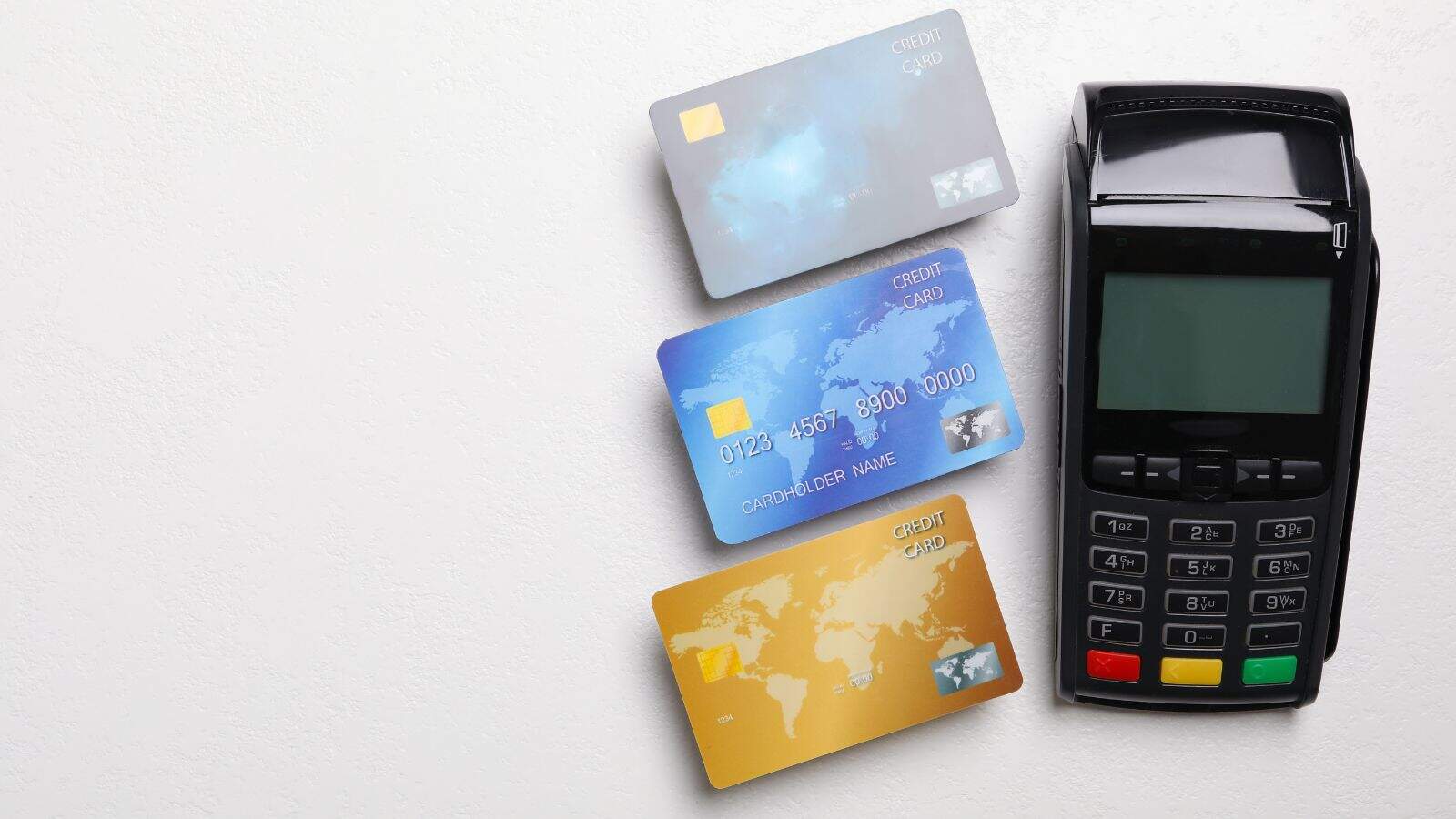 Three credit cards in gray, blue, and gold are arranged next to a black payment terminal on a white surface. The cards display generic names and numbers.