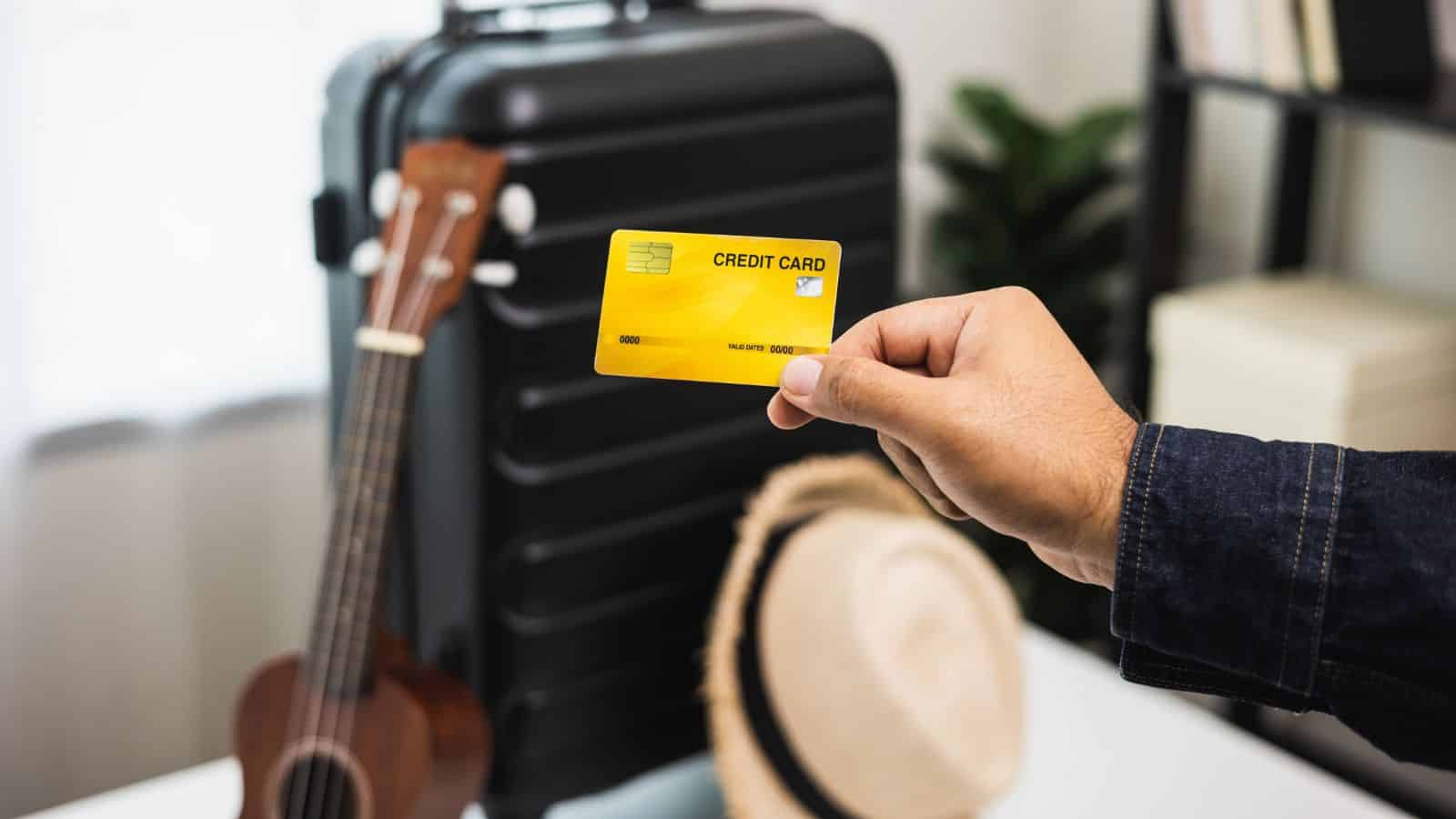 A person holds a yellow credit card in front of a black suitcase. Nearby, a straw hat with a black band and a small wooden ukulele are on a white surface. The background includes a plant and shelves.