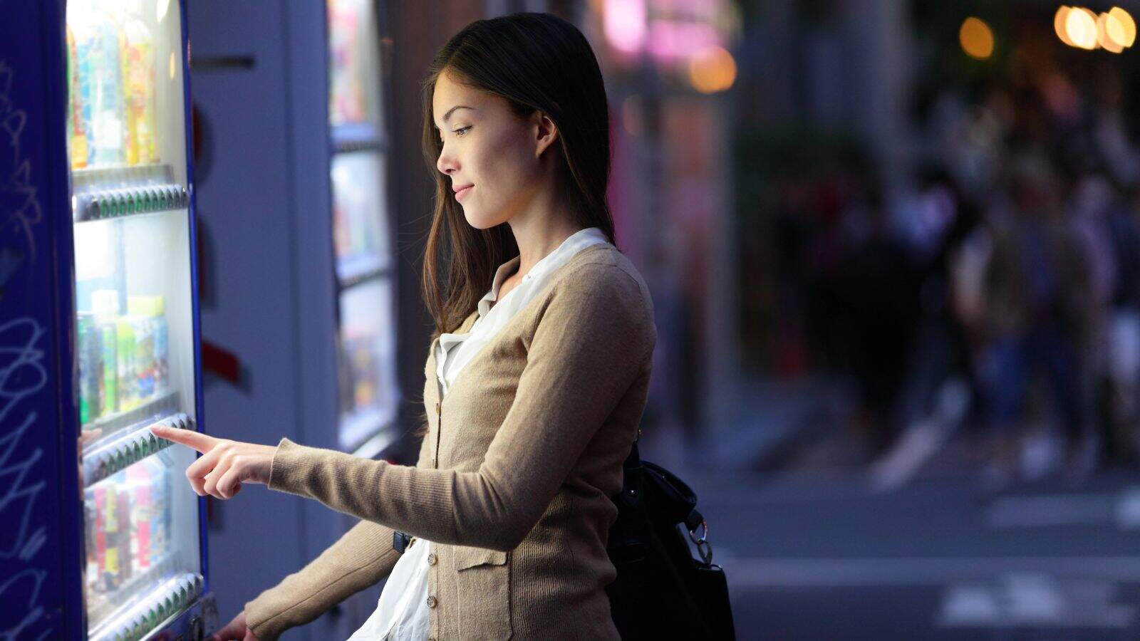 A woman stands outside at night, looking at a brightly lit vending machine and pressing a button to make a selection. She carries a black bag and wears a beige cardigan over a white shirt. The background is blurred.