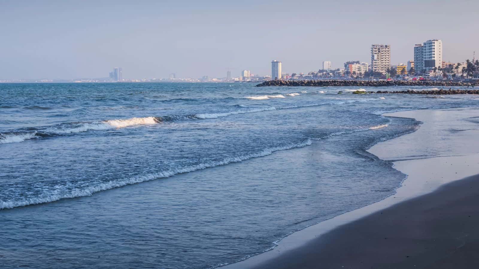 Waves gently reach the sandy shore under a clear sky, with several mid-rise buildings and palm trees visible along the distant coastline.