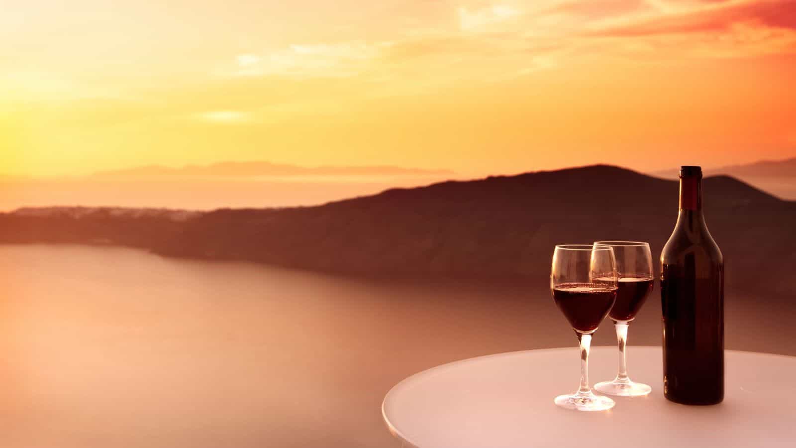 A bottle of red wine and two filled wine glasses sit on a round white table, overlooking a calm body of water and distant hills at sunset, with an orange and pink sky.