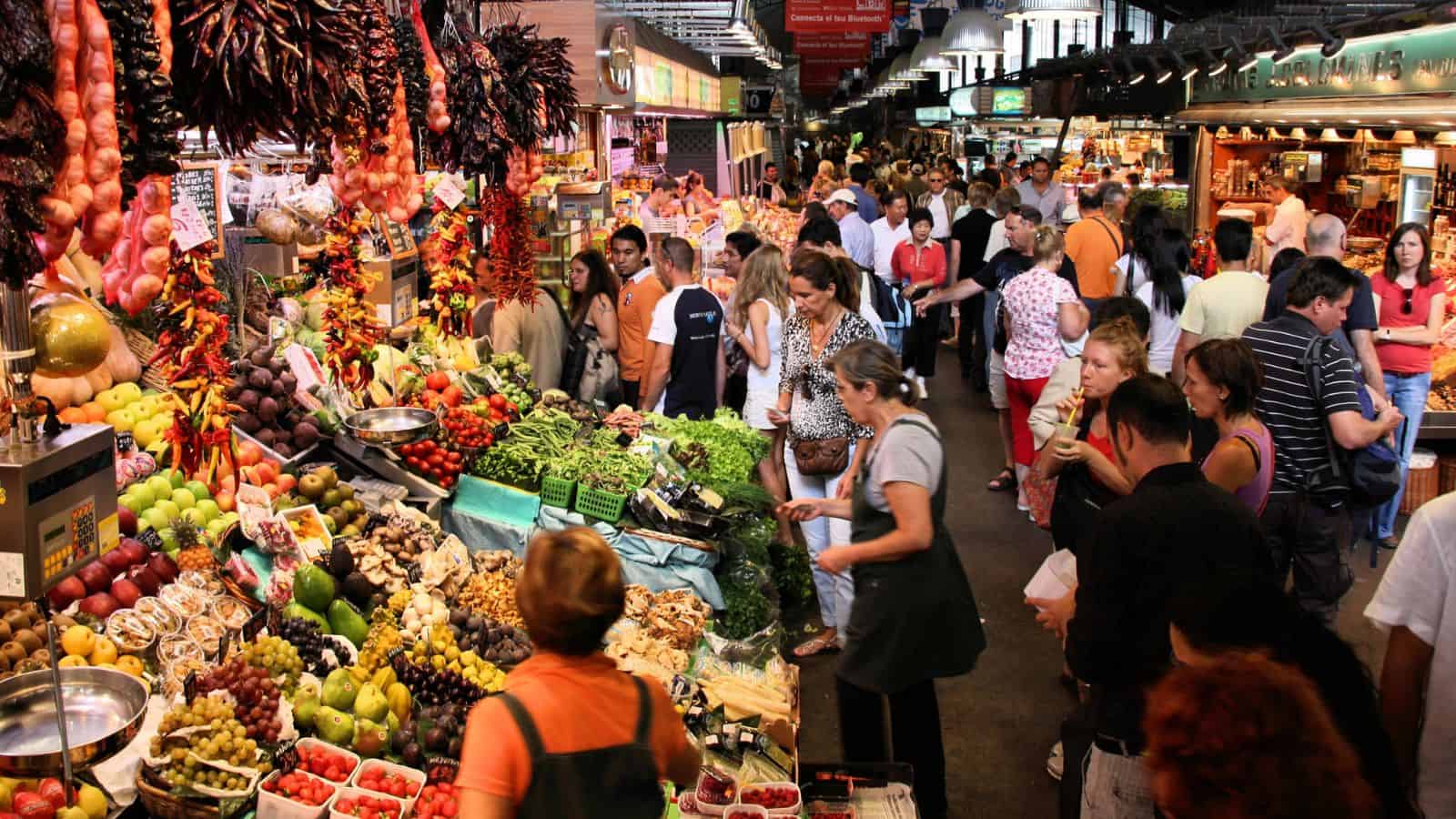 A busy indoor market with many people shopping for fresh fruits, vegetables, and other produce displayed on colorful, crowded stalls. Overhead lights illuminate the lively scene.