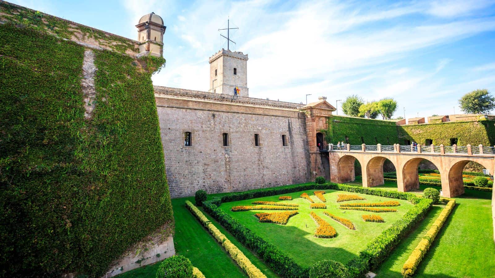 A stone fortress with green ivy-covered walls, a central tower, and arched bridge spans over a manicured garden featuring geometric patterns of trimmed hedges under a blue sky.