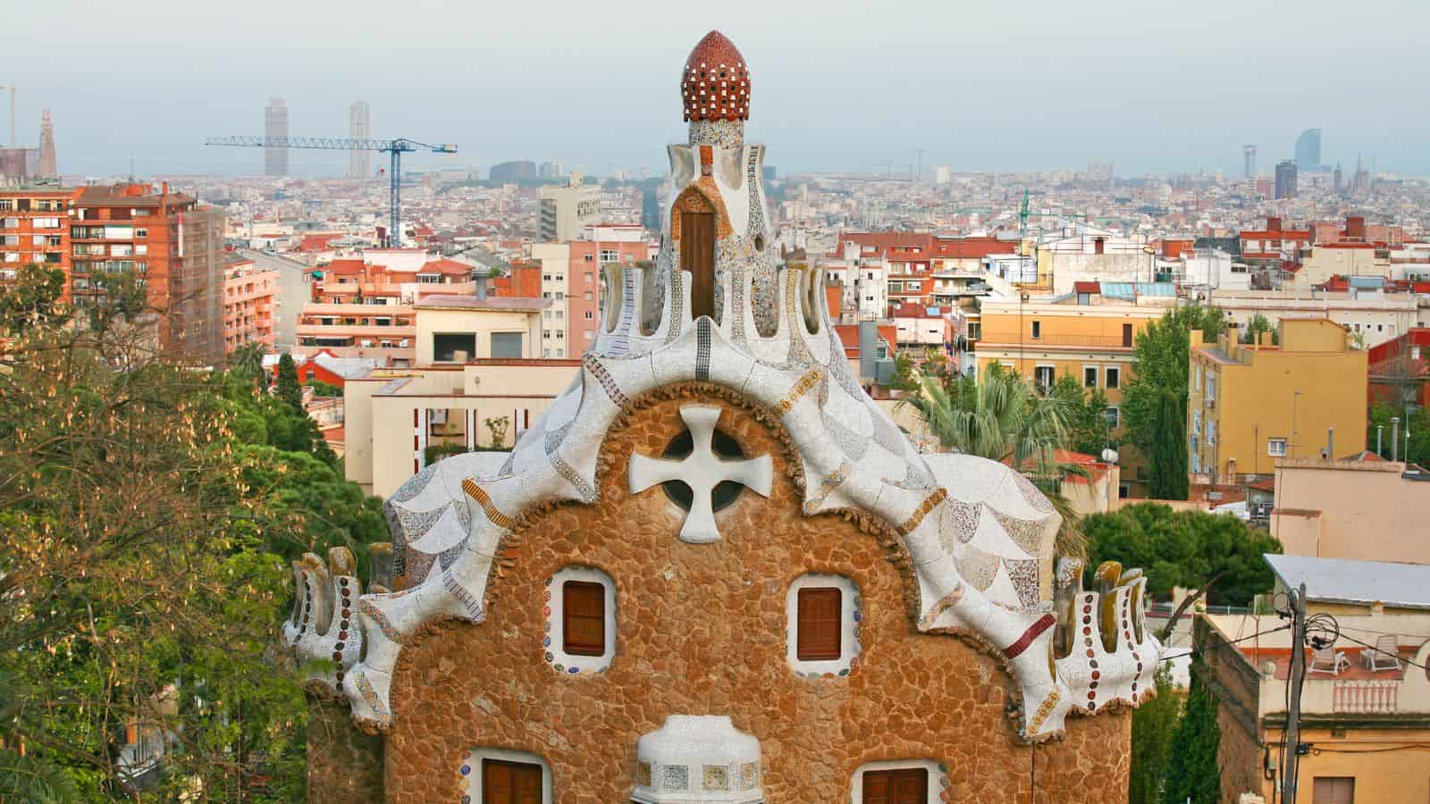 A stone building with white mosaic trim and a unique rooftop stands in the foreground, with an urban cityscape, various buildings, and distant skyscrapers visible in the background.