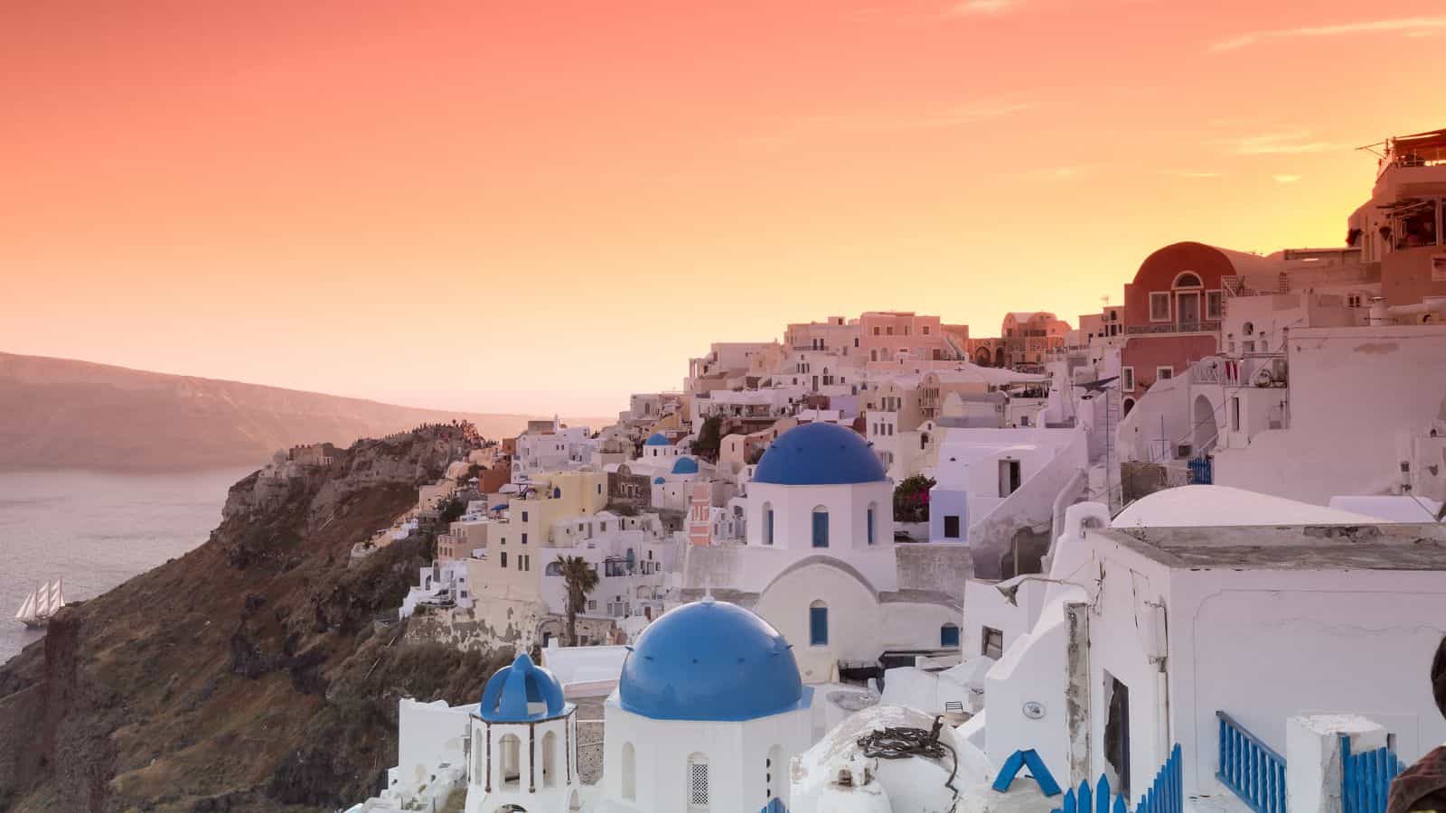 Buildings with white walls and blue domes are clustered on a hillside overlooking the sea at sunset. The sky is orange, and the architecture is typical of a coastal Mediterranean town.