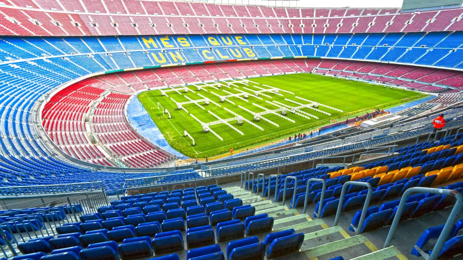 A wide-angle view of an empty football stadium with red and blue seats, yellow seats in one section, and the words "MÉS QUE UN CLUB" on one stand. The field has white lighting rigs placed on the grass.