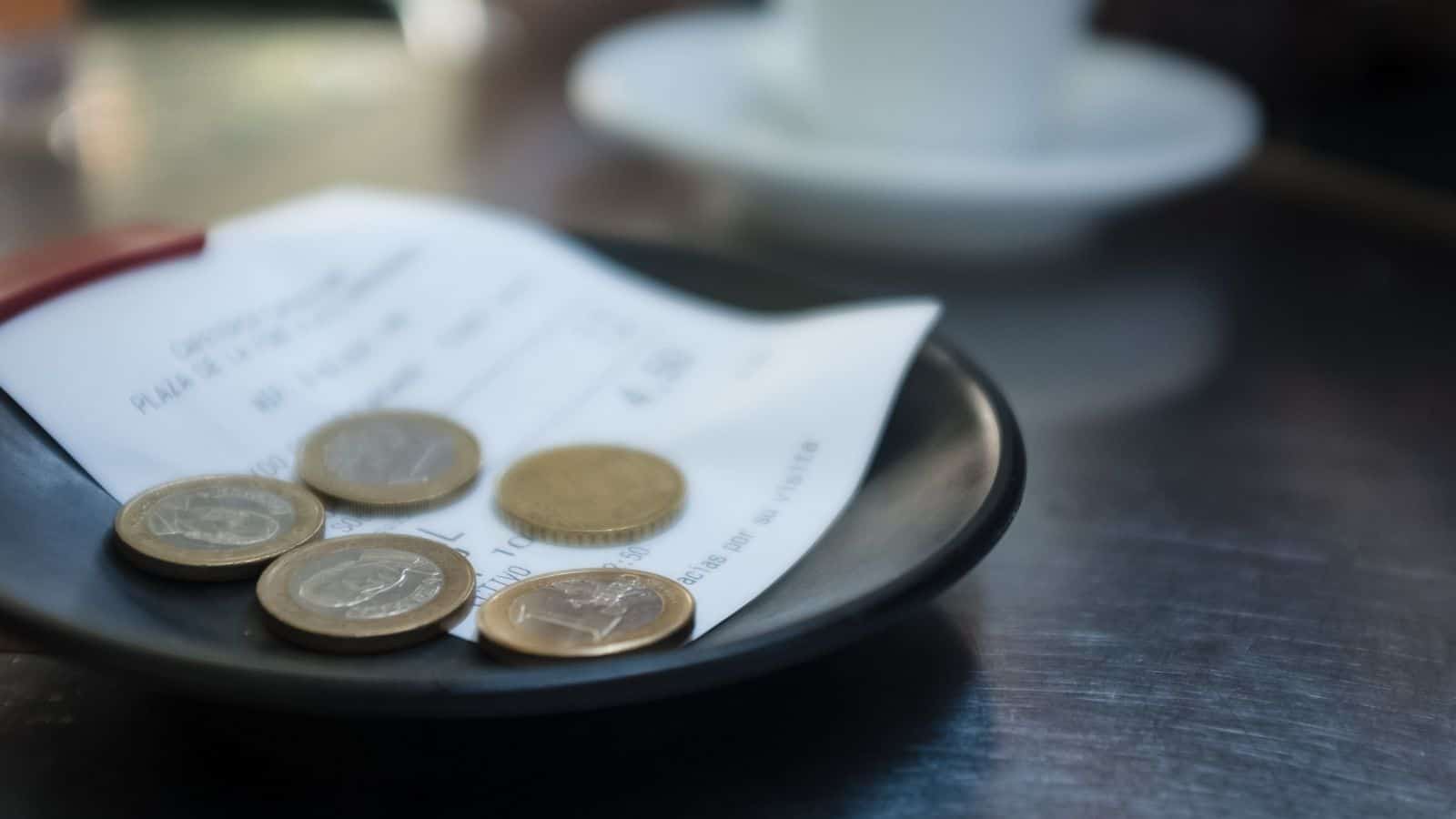 A close-up of a black plate with a restaurant receipt and several euro coins on it, placed on a metallic table with a white cup and saucer blurred in the background.