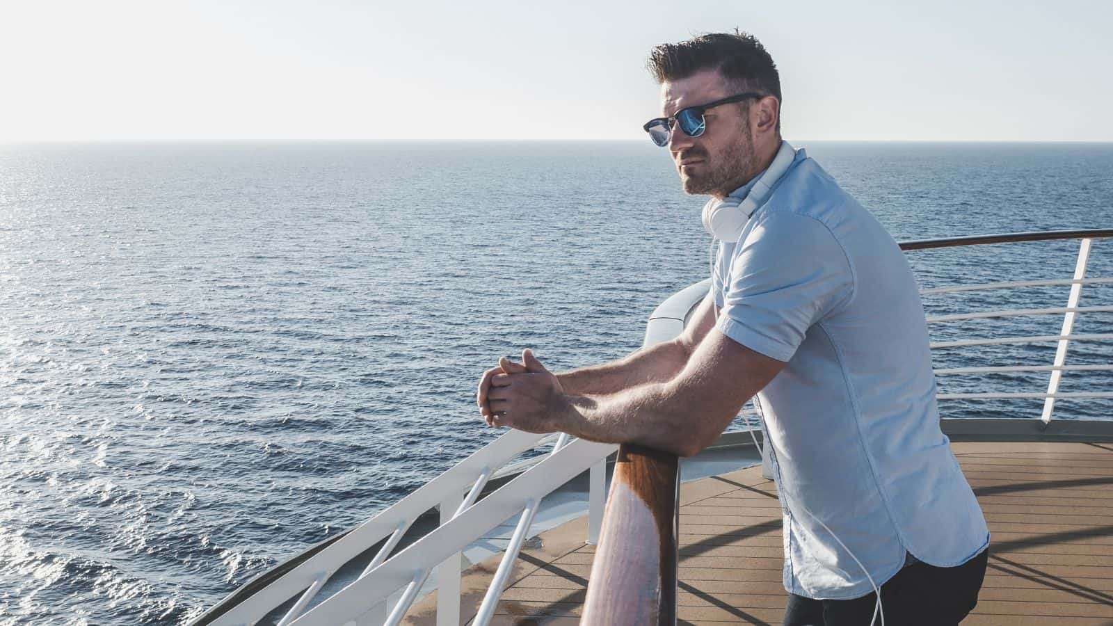 A man wearing sunglasses and headphones stands on the deck of a ship, leaning on the railing and looking out at the open sea under a clear sky.