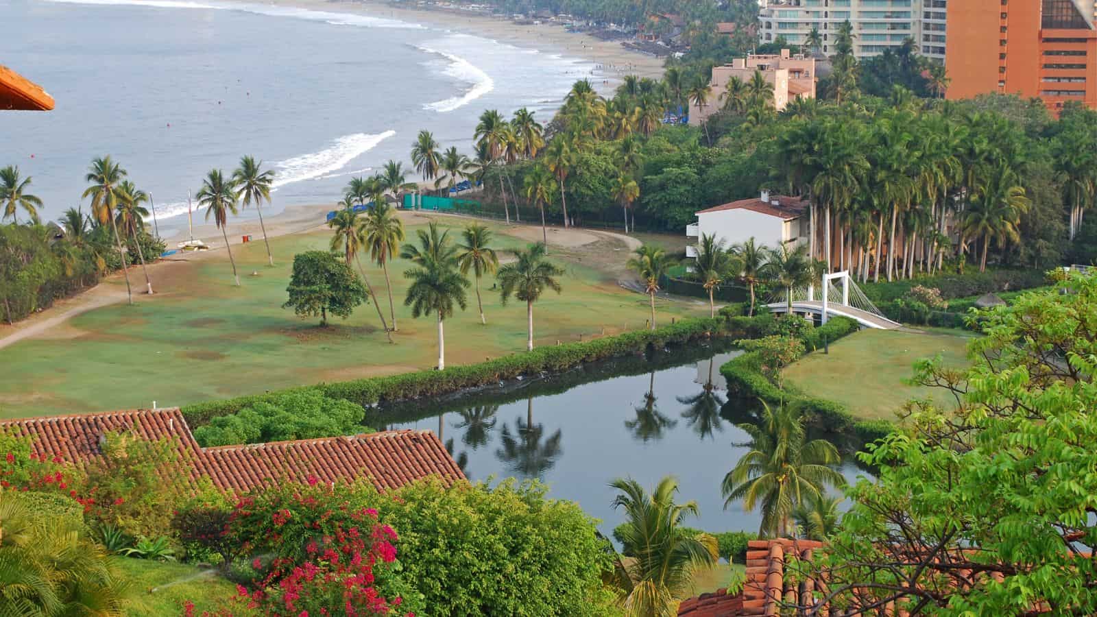 A coastal area with palm trees, a grassy landscape, a small pond with a white footbridge, and buildings near the shoreline. The ocean and sandy beach are visible in the background.
