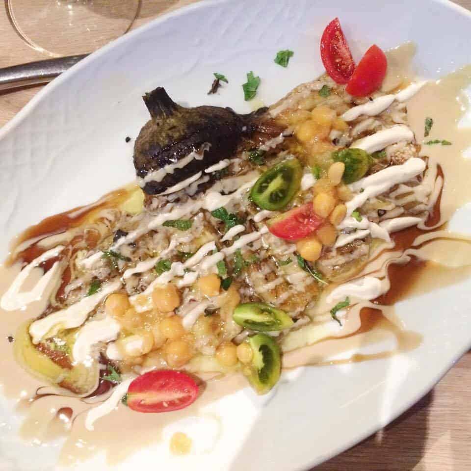 Grilled eggplant topped with sauce, herbs, cherry tomatoes, and yellow pearls on a white plate.