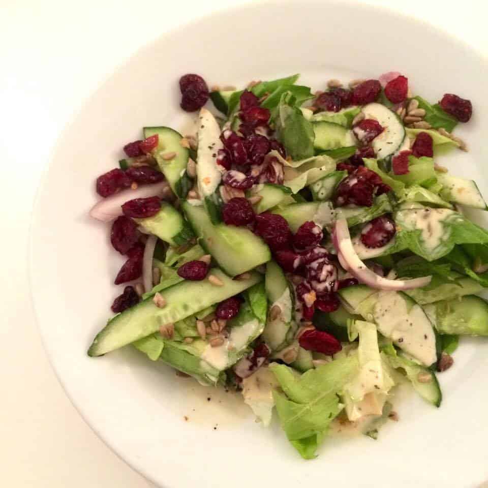 A fresh salad with mixed greens, cucumber, red onion, dried cranberries, seeds, and dressing in a white bowl.