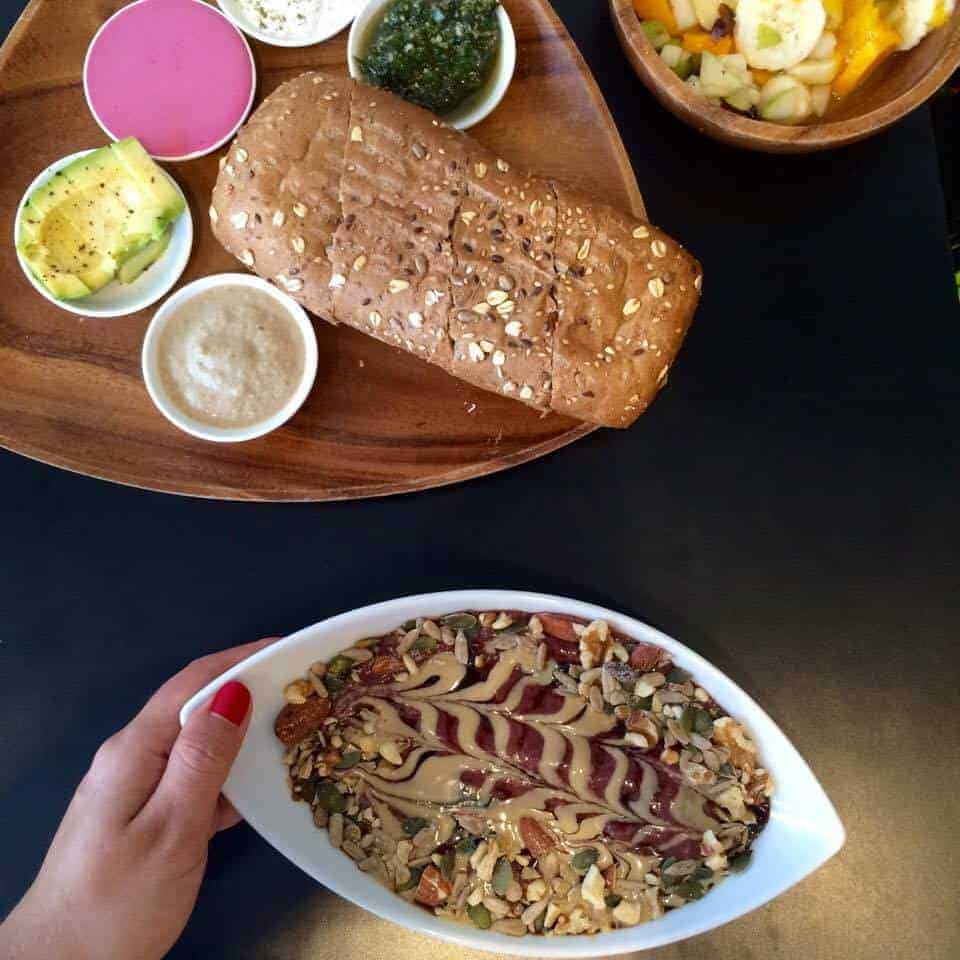 A hand holds a bowl of smoothie topped with seeds and nut butter; bread and spreads are on a wooden tray above.