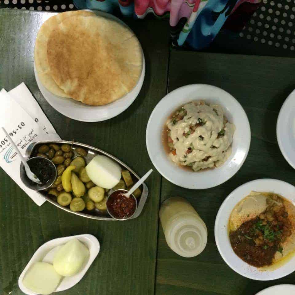 A table set with pita bread, hummus, pickles, onions, sauces, and a bottle of dressing.