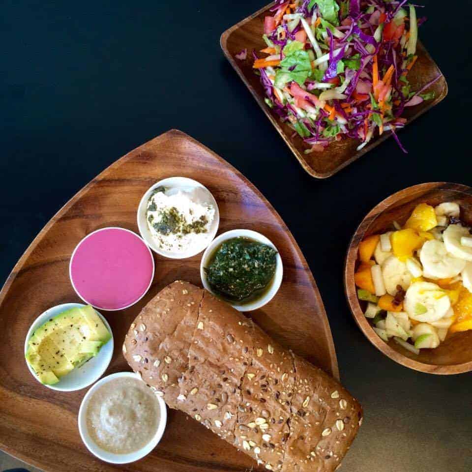 Triangular wooden plate with bread, dips, salad, and fruit bowl on a black table.