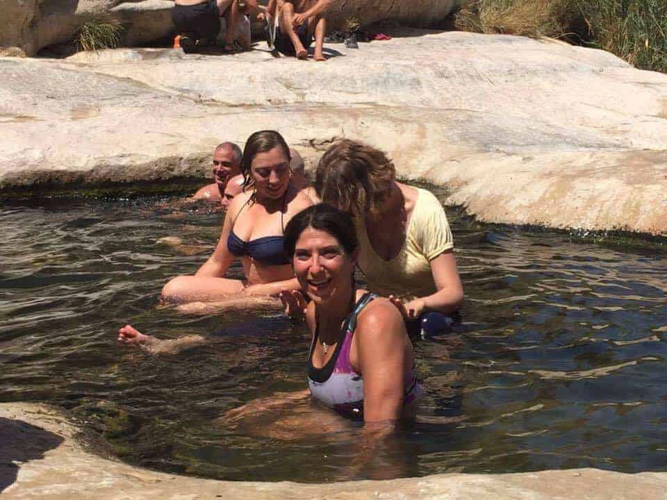 Three people relaxing in a rocky natural pool, with others sitting on rocks in the background.