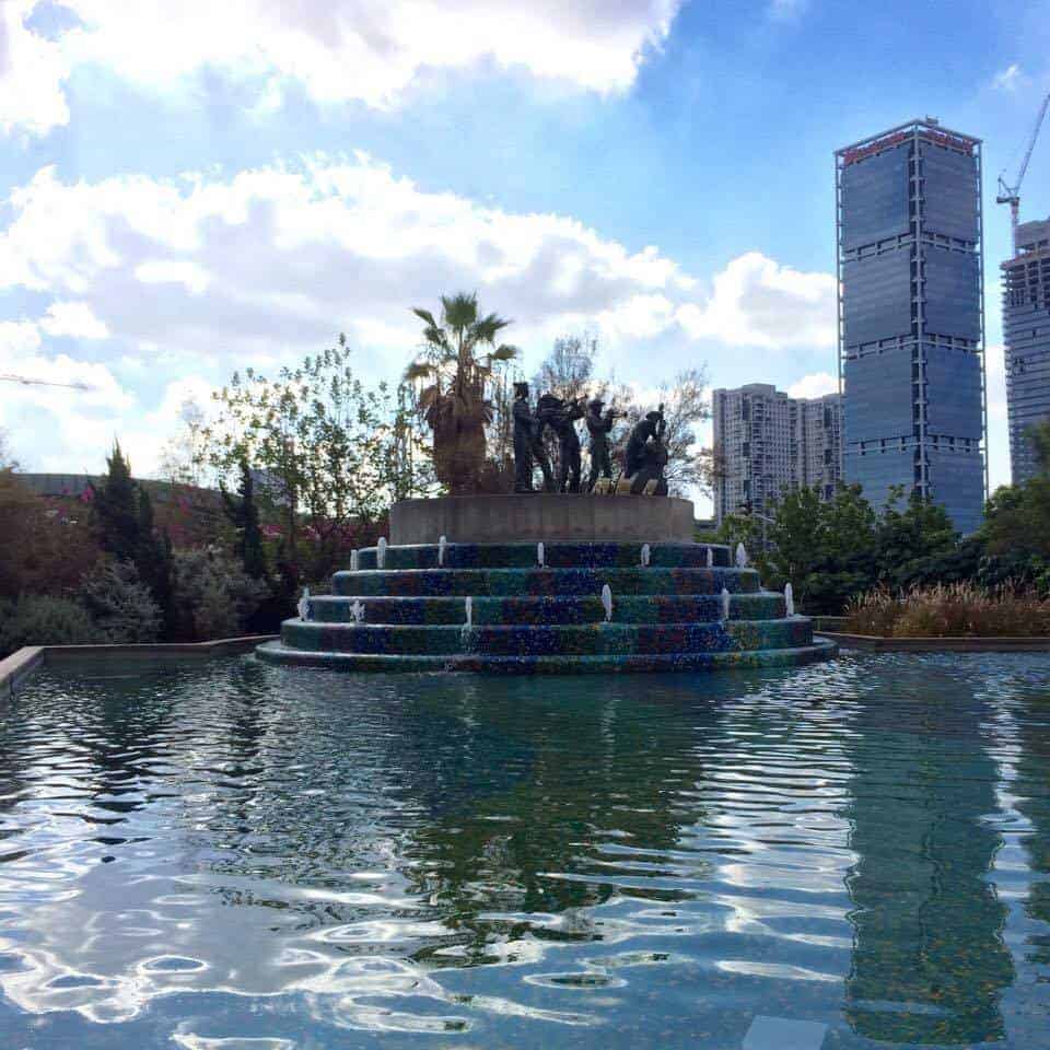 A tiered fountain with sculptures, surrounded by water, with modern buildings in the background.