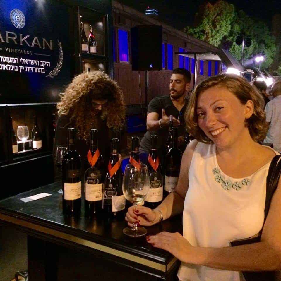 Woman smiling with a glass of wine at a nighttime wine tasting event, bottles displayed on the table.