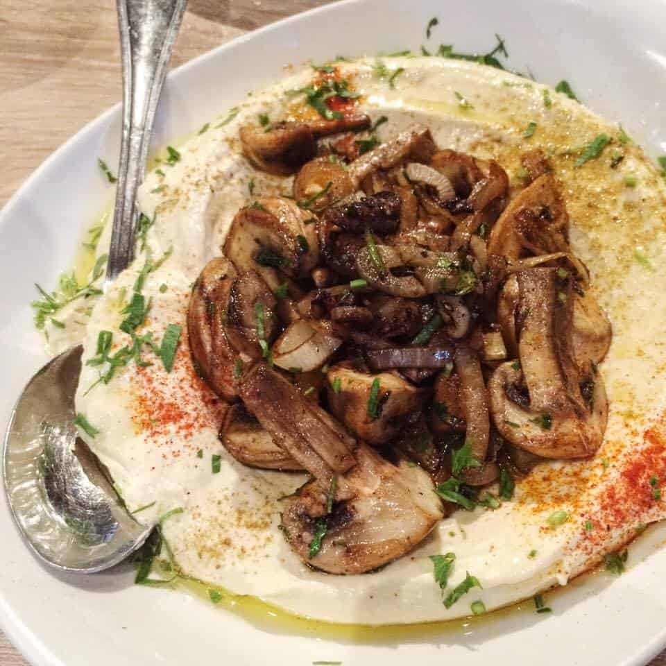 A plate of hummus topped with sautéed mushrooms, herbs, and spices, served with a spoon.