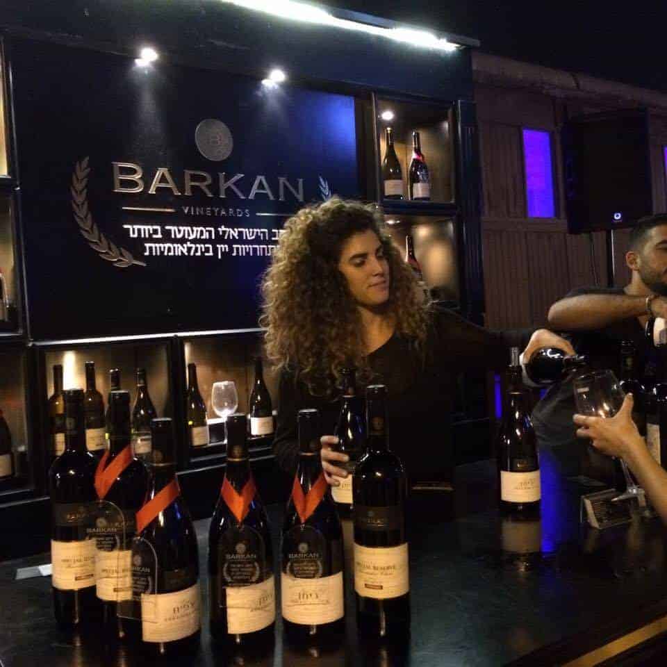 A woman pours wine at a Barkan Vineyards booth, surrounded by bottles on a counter at night.