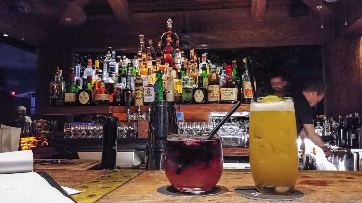 Two colorful cocktails on a bar counter with shelves of liquor bottles in the background.
