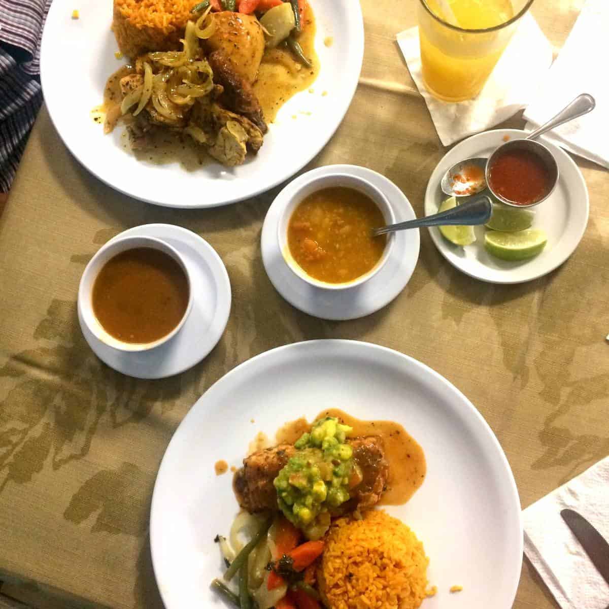 Two plates of rice, vegetables, chicken, soup bowls, sauces, lime wedges, and a glass of orange drink.