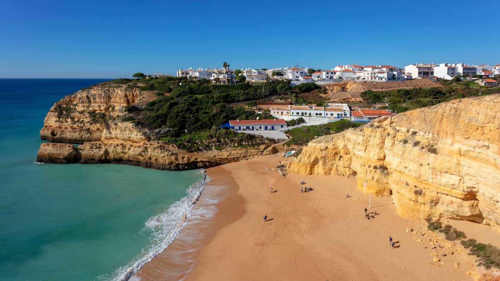 Golden sandy beach bordered by cliffs, turquoise sea, and white buildings on a hill under a clear blue sky.