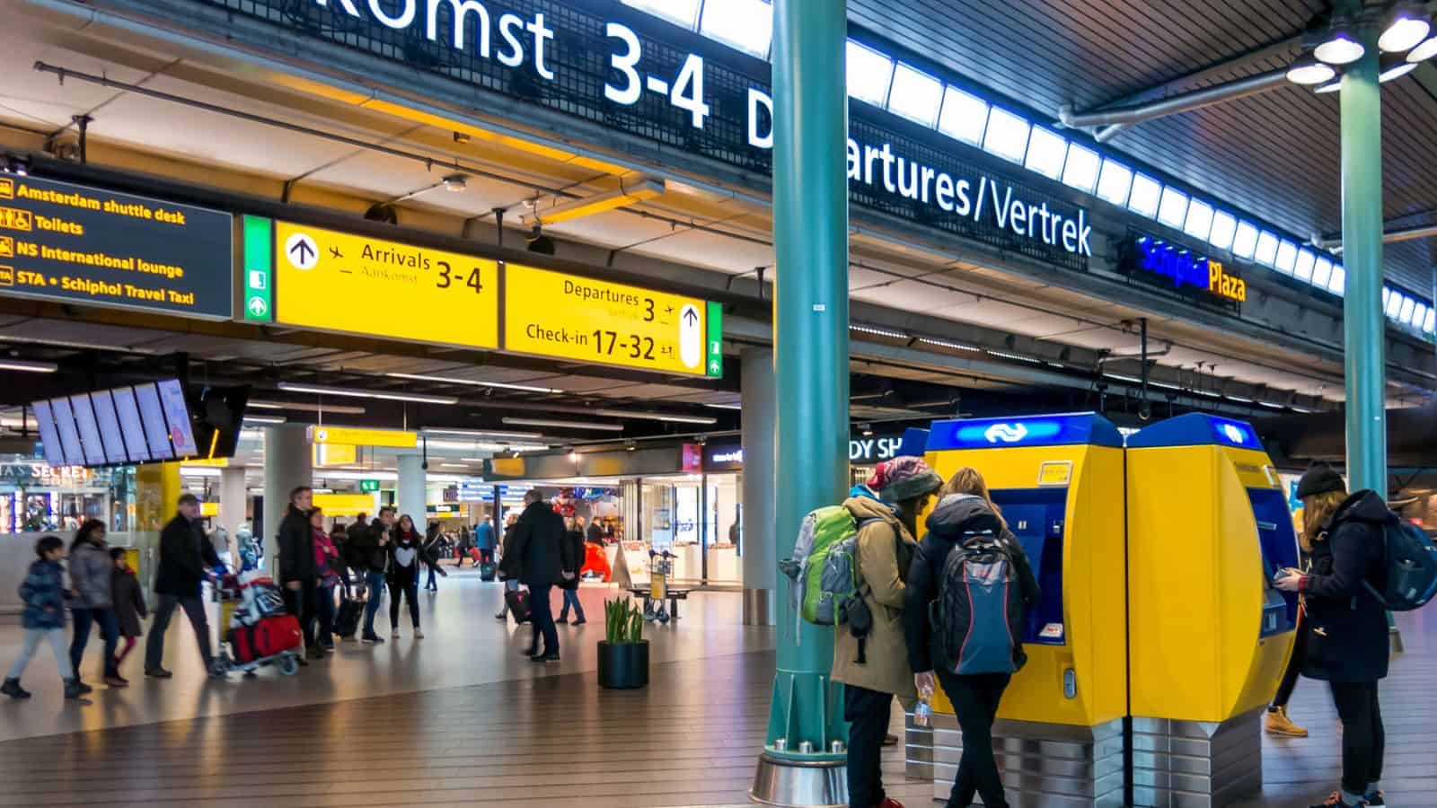 Passengers walk through a busy airport terminal with yellow signs overhead showing directions for arrivals, departures, and check-in; some people use a ticket machine on the right.