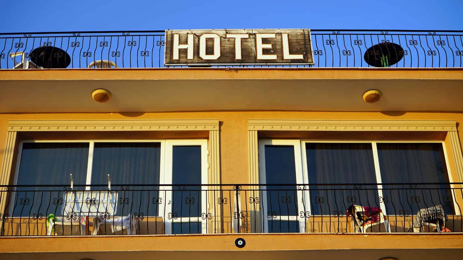 Hotel balcony with patio chairs, towels, and a large "HOTEL" sign above the railings under a clear blue sky.