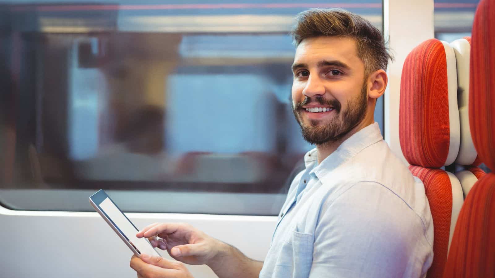 A man with a beard sits on a train ride next to a window, holding and using a tablet. He is wearing a light-colored shirt and looking at the camera, with red and white seats visible beside him—perfect posture for good train etiquette.