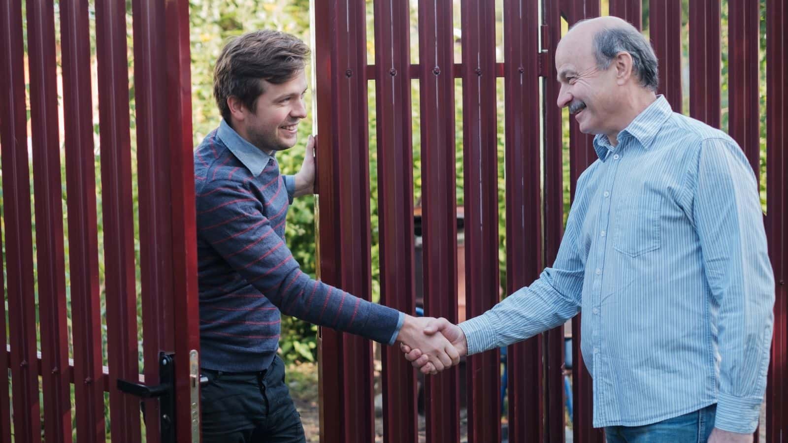 Two men smiling and shaking hands by a red metal fence outdoors.