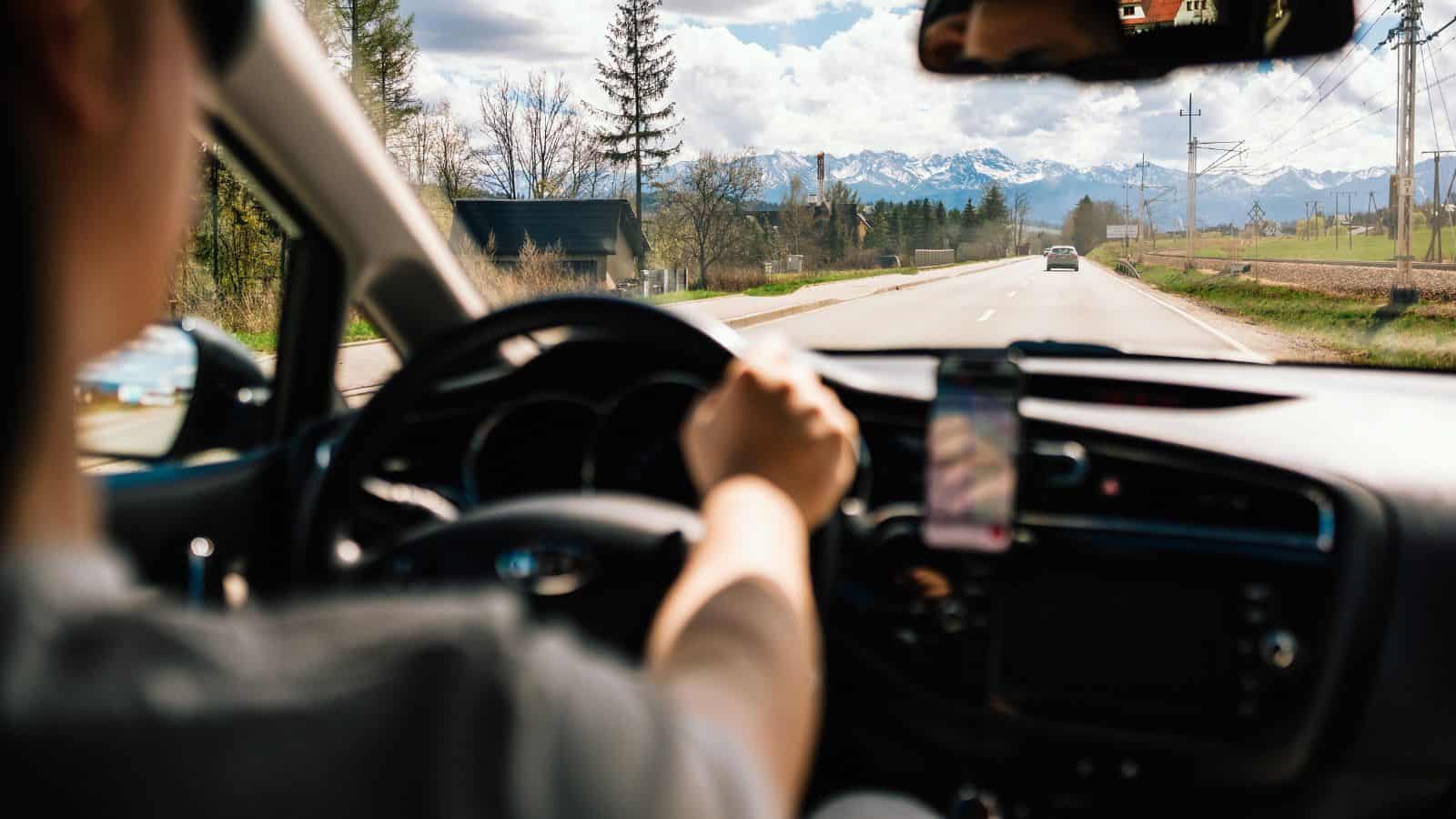 A person drives a car down a rural road with fields, trees, and mountains visible ahead. The dashboard, steering wheel, and a phone mounted on the console are in view. It is daytime with partly cloudy skies.