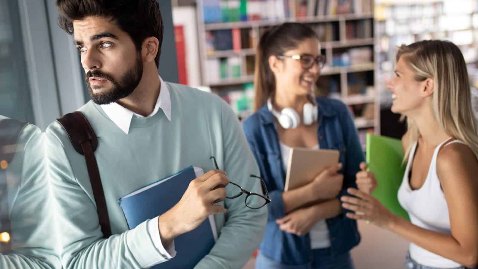 A serious man stands apart from two women talking and laughing in a library or study area.