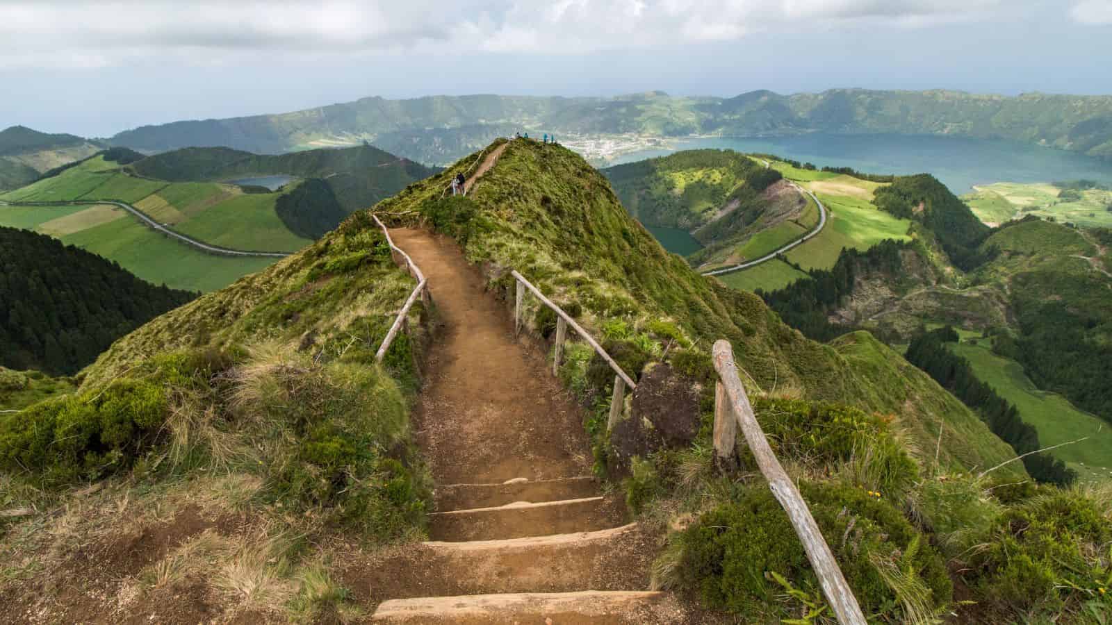 A dirt path with wooden railings leads up a green hill, overlooking lakes, fields, and mountains in the distance.