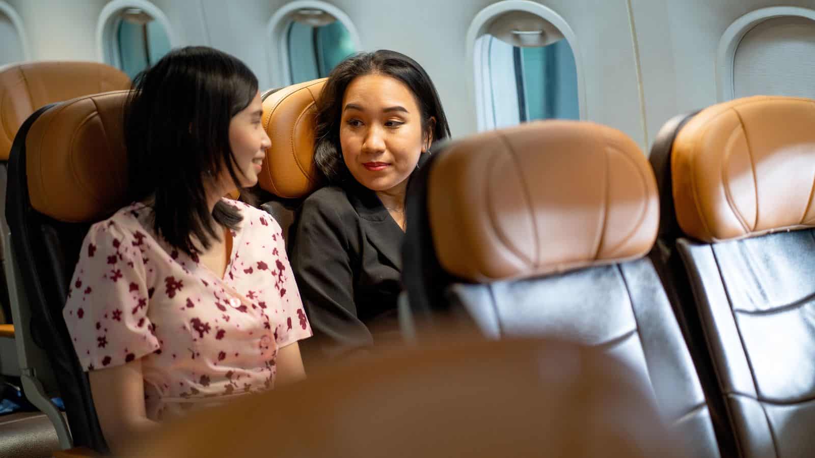 Two women are sitting next to each other on an airplane, having a conversation. Both are facing each other and smiling slightly, with empty seats and airplane windows visible in the background.
