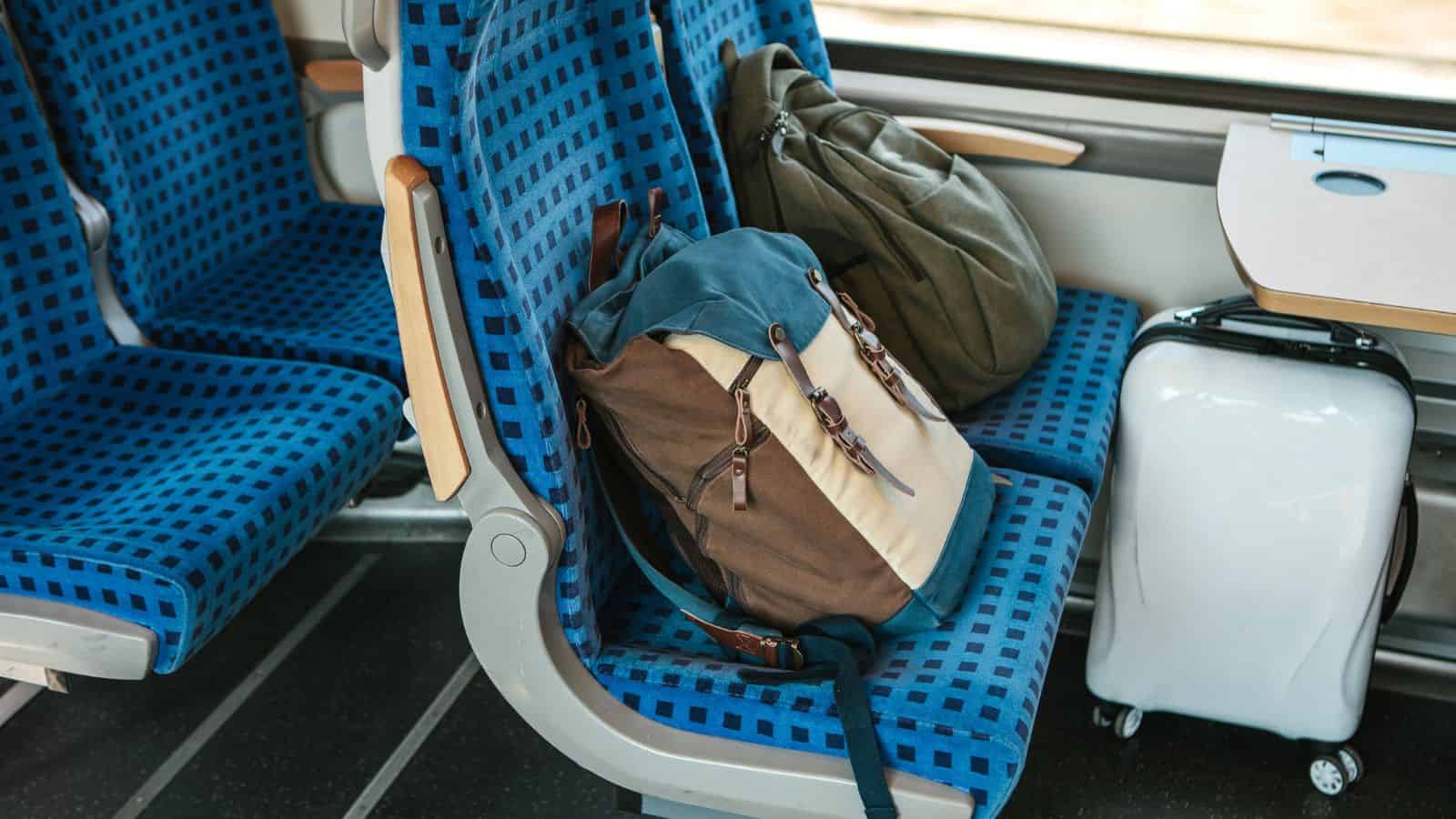 A blue-checkered train seat holds a brown and beige backpack and a green duffel bag. A small white carry-on suitcase stands on the floor beside the seat, following good train etiquette for an organized train ride.