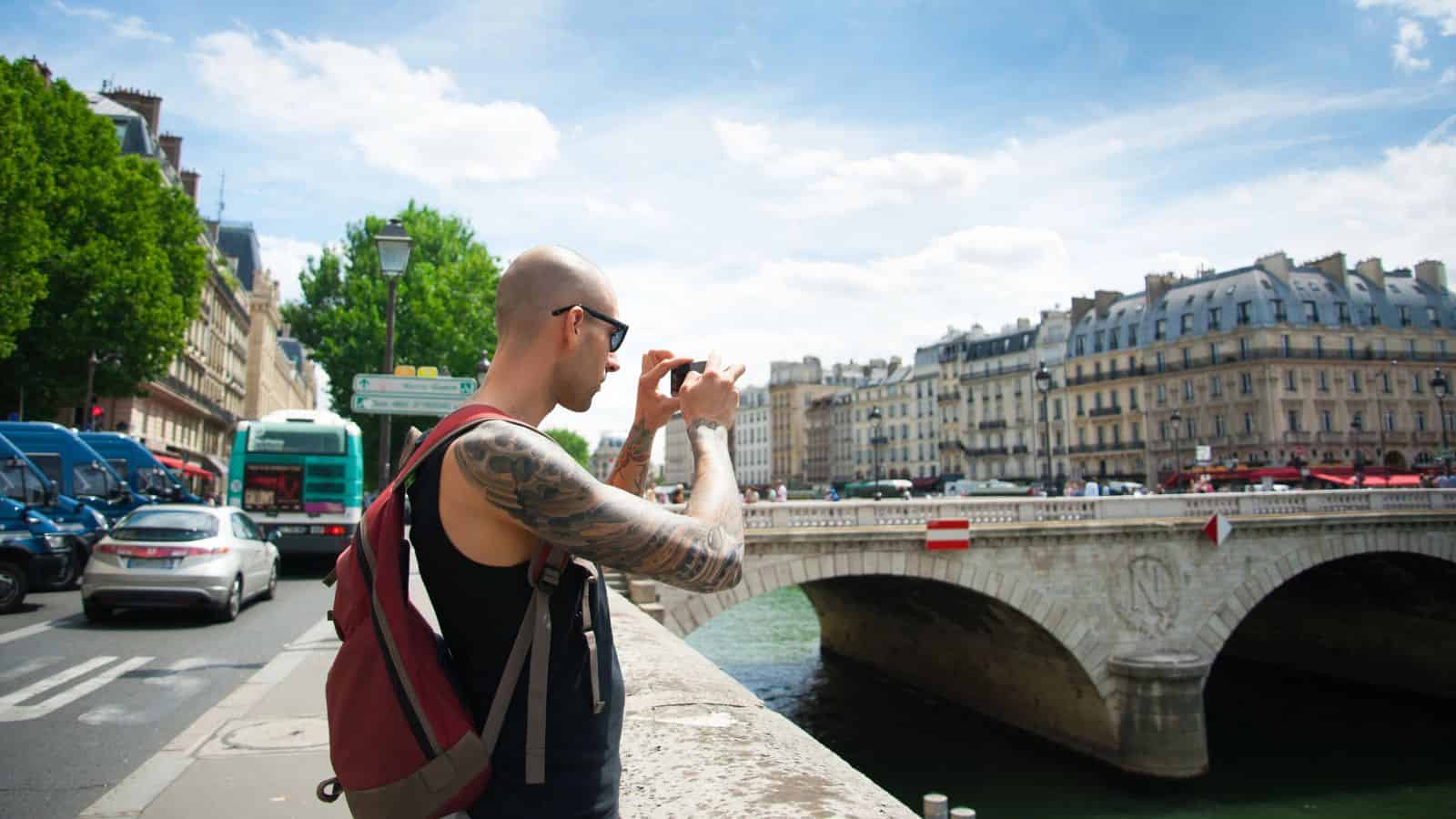 A man with tattoos takes a photo by a river and bridge in Paris, historic buildings behind, blending in seamlessly with locals and avoiding tourist mistakes.
