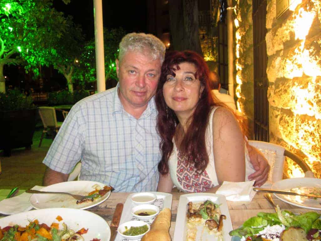A smiling couple sits together at an outdoor restaurant table with food and drinks.