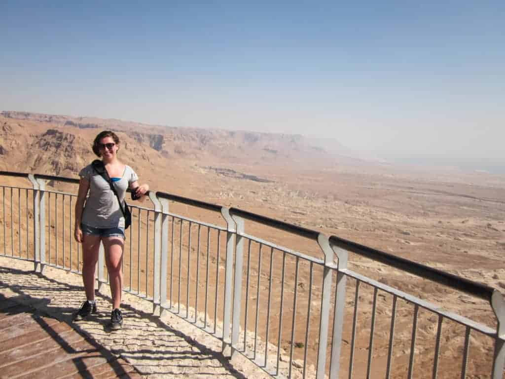 Woman in shorts and sunglasses stands on a lookout platform overlooking a vast desert landscape.