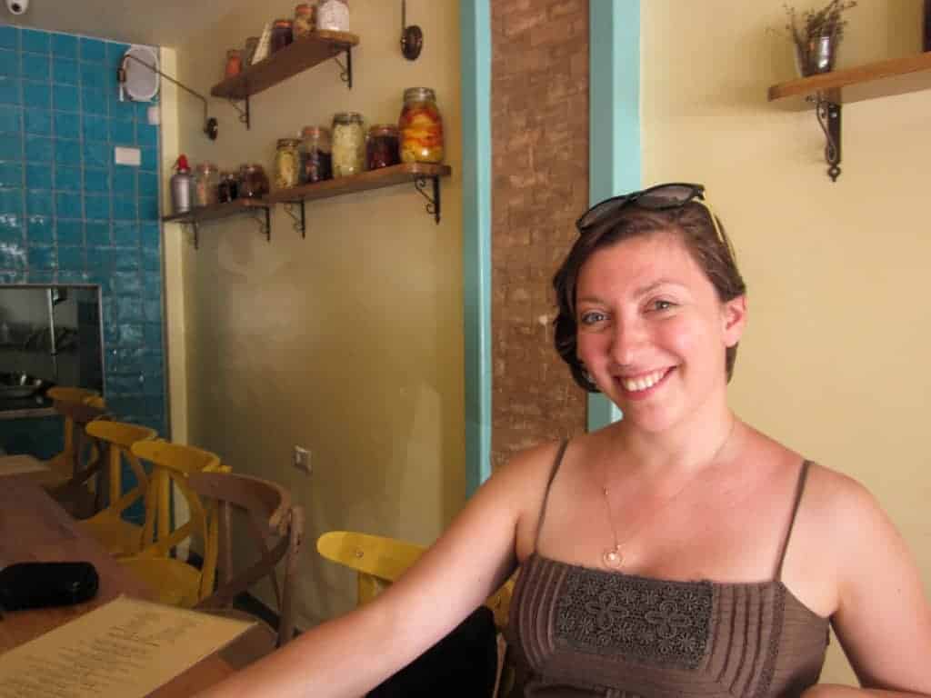 A smiling woman sits at a table in a cozy café with jars on shelves and blue tiled walls.