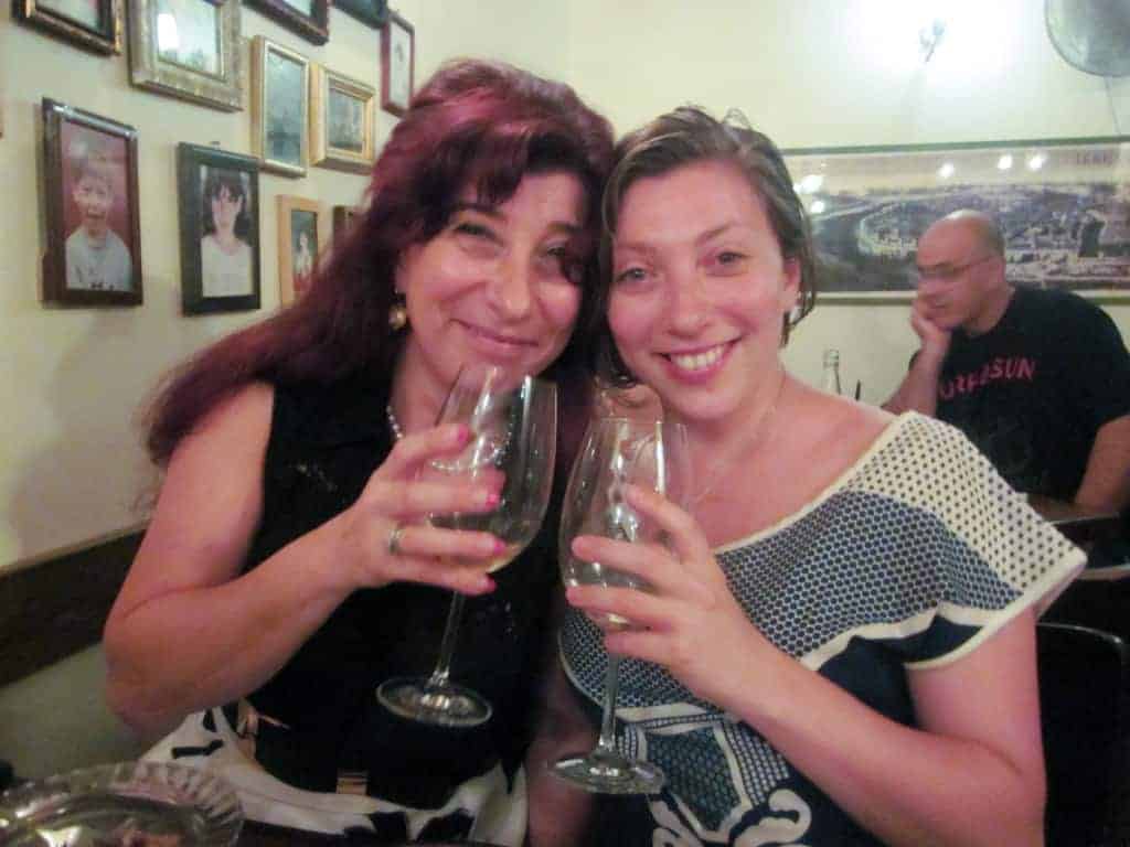 Two women smiling and holding wine glasses in a cozy restaurant with framed photos on the wall behind them.