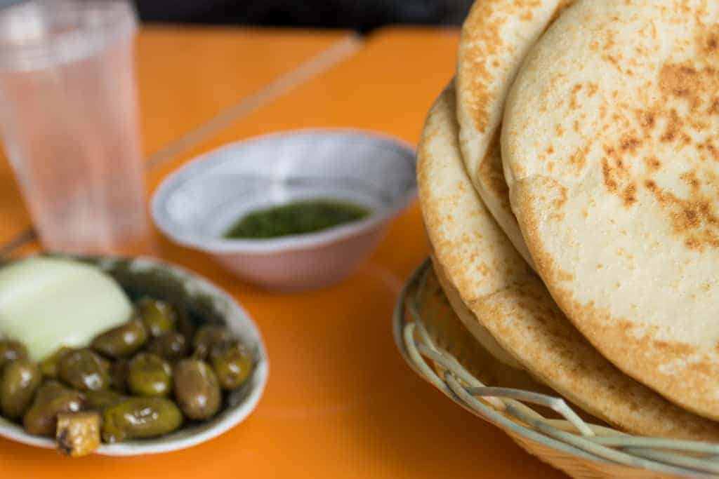 A basket of flatbreads with a side of olives, onions, and a small bowl of green sauce on an orange table.