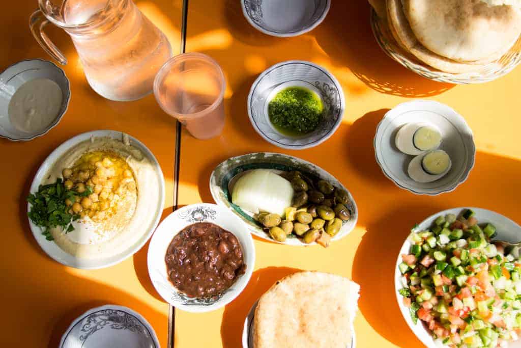 A table with hummus, pita, salad, olives, eggs, beans, sauces, and a jug of water on a yellow surface.