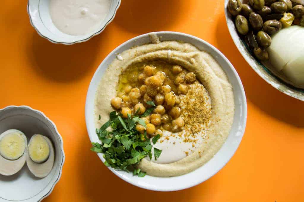 Bowl of hummus topped with chickpeas, herbs, and oil, surrounded by eggs, olives, and sauces on an orange table.